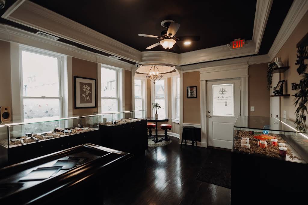 Interior view of a modern cannabis dispensary featuring display cases with products, stylish seating, and large windows allowing natural light. The decor includes a ceiling fan and decorative elements, creating a welcoming atmosphere for customers.