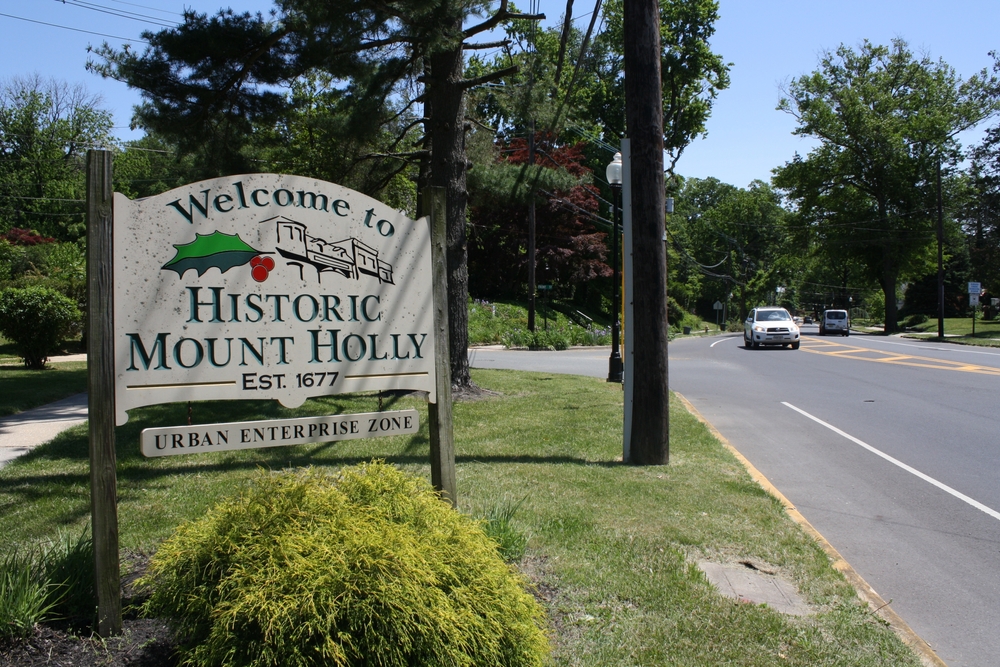 Welcome sign for Historic Mount Holly, established in 1677, located in an Urban Enterprise Zone, surrounded by greenery and roads.